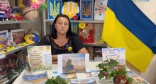 Учасниця акції сидить за столом, на якому розміщені патріотичні плакати та прапор України