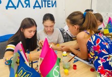 Діти малюють фарбами за столом, зосереджено працюючи над спільною роботою, поряд стоять прапорці України та Бахмутської громади.