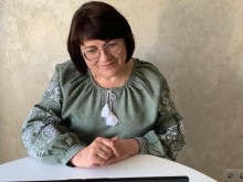 Жінка в окулярах та зеленій вишиванці сидить за білим столом, зосереджено дивлячись у монітор ноутбука.
