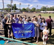 Група людей стоїть на стадіоні, де відбуваються мотоциклетні перегони та тримають прапор з надписом: гуманітарний хаб "Єднання"