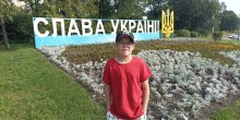 Хлопчик в окулярах, червоній футболці та бейсболці стоїть на тлі клумби та великого напису «Слава Україні!».