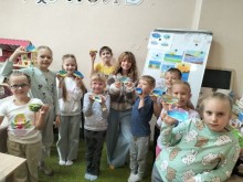 Група усміхнених дітей з гордістю демонструє свої барвисті малюнки в класі. На дошці в задньому плані виставлено ще більше художніх робіт.