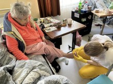 Літня жінка сидить на ліжкуу прихистку. Лікар подолог проводить огляд пальців.