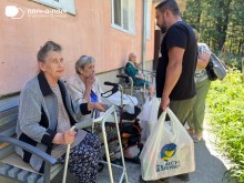 Жінки похилого віку сидять на вулиці біля будівлі, поруч з ними ходунки. Заступник начальника Часовоярської громади стоїть біля них з пакетом гуманітарної допомоги. Спілкуються.