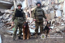 Двоє кінологів та службові собаки на місці влучання. Позаду зруйнована будівля.
