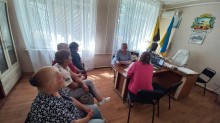 Кілька людей сидять обличчям до жінки за столом, яка, проводить онлайн-зустрічі з ветеранами та членами родин загиблих захисників і захисниць України. На столі видно ноутбук, що відображає відеодзвінок, а на стіні за столом висить український прапор.