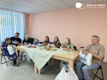 Жінки та чоловік похилого віку сидять за столом. Поруч пакети з гуманітарною допомогою. На столі лежать книги та журнали. З ними спілкується Руслан Прийменко
