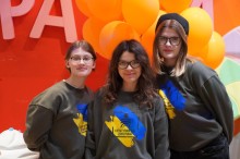 Три дівчини в однакових оливкових світшотах з написом «Культурний хаб Донеччина» на фоні помаранчевих кульок.