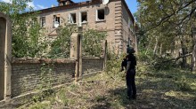 Правоохоронець стоїть біля двоповерхового будинку, в якому обстрілом вибило вікна.