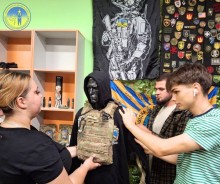 Молода дічина та двоє хлопців розглядають одягнений на манекен бронежилет зеленого кольору.