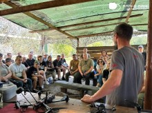 Чоловіки та жінки сидять на  лавочках. Інструктор стоїть спиною до камери. Поруч на столі лежать дрони.