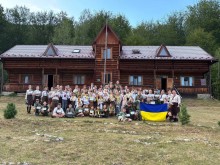 Учасники пластового табору «Земля велетнів» тримають український прапор перед дерев'яним будинком.