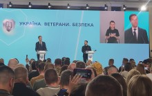 На сцені двоє чоловіків стоять за трибунами, звернених до аудиторії. На великому екрані за ними зображені ці ж люди, один з яких — Президент України Володимир Зеленський, а інший — Генеральний секретар НАТО Марк Рютте. Збоку на екрані також видно жінку-сурдоперекладача.