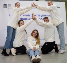 П’ятеро молодих людей у білих худі утворюють фігуру у формі будинку, тримаючи долоні разом над головою дівчини, яка сидить посередині, під час молодіжного заходу для молоді Донеччині.