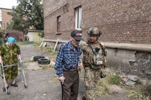 Поліцейський допомагає літньому чоловіку евакуюватись з міста Костянтинівка. Веде чоловіка під руку. Позаду літня жінка з ходунками