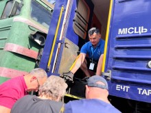 Працівник з благодійної організації в вагоні потяга допомагає маломобільній жінці піднятись до вагона. Поруч чоловіки, які допомагають.