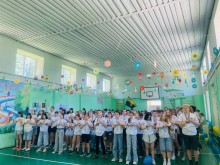 Діти в спортивній залі виконують рухи флешмобу з руками на серці, навколо - паперові квіти й прапори, панує святкова атмосфера.
