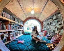 Дівчина сидить у затишному кімнатному просторі з круглим вікном, оточеному книжковими полицями та декоративними подушками, читаючи книгу.