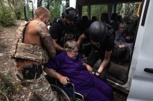 Евакуація. Волонтер та співробітники поліції у бронежилетах заносять літню жінку, яка знаходиться в інвалідному візку до броньованого автомобіля, де знаходиться ще одна літня жінка.