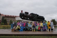 Учасники пластового табору «Земля велетнів» у дощовиках під дощем, стоять біля експоната старого паротяга