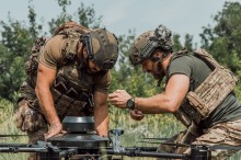 Двоє солдатів у тактичному спорядженні зосереджено працюють над великим дроном на відкритому повітрі. Вони зосереджені, що свідчить про командну роботу та точність. На задньому плані розмиті дерева.