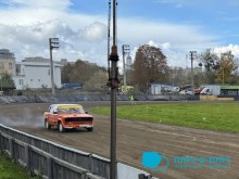 Червоно-жовтий гоночний автомобіль мчить по грунтовій дорозі, піднімаючи хмари пилу. Похмуре небо, дерева та промислові будівлі на задньому плані створюють динамічну атмосферу.