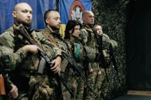 П'ять солдатів у камуфляжній формі стоять у строю, тримаючи гвинтівки. За ними на стіні висить синьо-жовтий прапор. Атмосфера серйозна.