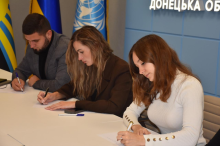 Працівники апарату облдержадміністрації сидять за столом і пишуть диктант на тлі прапорів та напису «Донецька область».