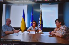 Учасники наради, чоловік та дві жінки, сидять за столом ,позаду учасниці прапори.