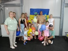 Дорослі й діти разом, тримають різнокольорові повітряні кульки на тлі синьо-жовтого прапора. Панує радісна атмосфера єдності.