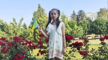 Маленька дівчинка з українським прапором на тлі квітучих троянд та зелені декламує вірш до Дня Конституції України.