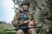 Боєць у повній екіпіровці тримає на руках рудого маленького кота. Збоку них дерева позаду поле.