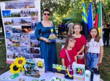 Жінка тримає сувенірну стелу Донеччини, поруч дівчата. Попереду стіл з сувенірною продукцією Донеччини.