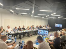 Всі учасники сидять за столами і дивляться екран, де виступають учасники, які приєднались онлайн.