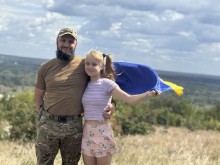 Дівчина з батьком-військовим на тлі природи. За спинами у обох розвивається синьо-жовтий прапор.