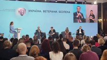 Зображено панельну дискусію. На сцені за столом сидять п'ять осіб, звернених до аудиторії, а зліва стоїть жінка, що виступає з промовою. На великому екрані за ними показано двох учасників дискусії.