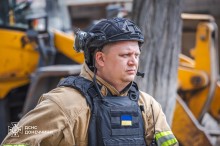 Рятувальник у повній екіпіровці, на бронежилеті наліпка прапора України. На задньому плані техніка.