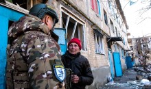 Солдат у камуфляжі розмовляє з літньою жінкою в червоному капелюсі біля пошкодженого житлового будинку. Ця сцена передає напругу і занепокоєння.