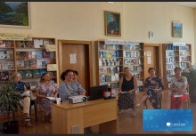 Читальний зал бібліотеки. Учасниці марафону сидять на стільцях, інші за столами. На столі стоїть проєктор. Позаду учасниць розташовані полиці з книжками.