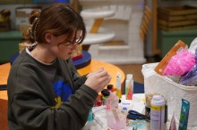 Дівчина в окулярах працює над поробом за столом, заставленим матеріалами для творчості.