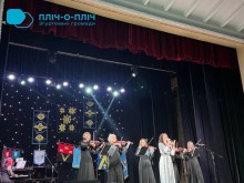 Дія на сцені. Жінки у чорних сукнях тримають скрипки та грають. Жінка у сірому платті співає у мікрофон.