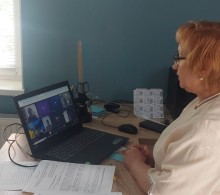 Заступниця начальника Іллінівської сільської військової адміністрації Наталія Лебедева, яка сидить за столом і дивиться в ноутбук, на екрані якого видно кілька учасників відеодзвінка. Перед жінкою на столі лежать документи.