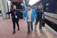 Волонтери ведуть чоловіка старшого віку на вокзаді