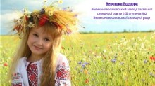 Дівчинка в українській вишиванці та вінку з польових квітів посміхається, стоячи на тлі квітучого поля.