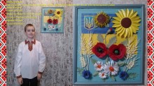 Хлопчик у вишиванці стоїть біля панно з українським прапором, соняшником, маками, ромашками та пшеницею, яку зробив власноруч.