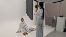 Фотограф знімає жінку в традиційному білому вбранні з червоною вишивкою, яка сидить на підлозі