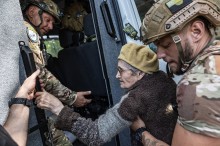 Літня жінка в окулярах однієї рукою тримається за поручень автомобіля. Поліцейські допомагають.