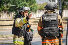 Двоє співробітників ДСНС, стоять на вулиці. Один із чоловіків рукою показує у бік, інший стоїть спиною, в нього на бронежилеті написано ДСНС. Обидва у захисних костюмах.