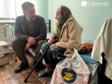 Заступник начальника Часовоярської міської військової адміністрації Руслан Прийменко сидить поруч біля жінки похилого віку, спілкується, поруч стоїть пакет гуманітарної допомоги.