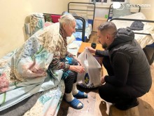 Жінка сидить на кроваті вкрита ковдрою біля неї Заступник начальника Часовоярської міської військової адміністрації Руслан Прийменко, сидить навколішках та передає пакети з гуманітарно допомогою.